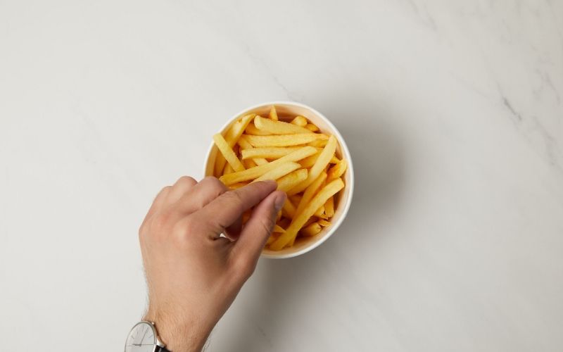 Männliche Hand mit Armbanduhr greift eine einzelne Pommes aus einer kleinen weißen Schale in der weitere Pommes liegen.