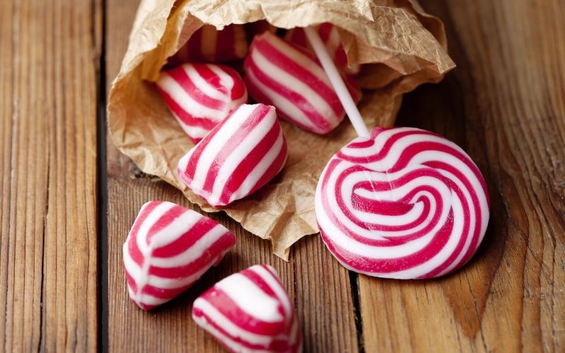 Süßigkeiten in rot und weiß gestreift liegen in einer Papiertüte auf einem Holztisch. Einige der Bonbons liegen auf dem Tisch vor der Tüte.