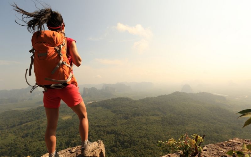 Dunkelhaarige Frau mit Rucksack steht am Felsrand auf einem Berg und schaut in die Weite. Wälder und Gebirge umgeben sie.