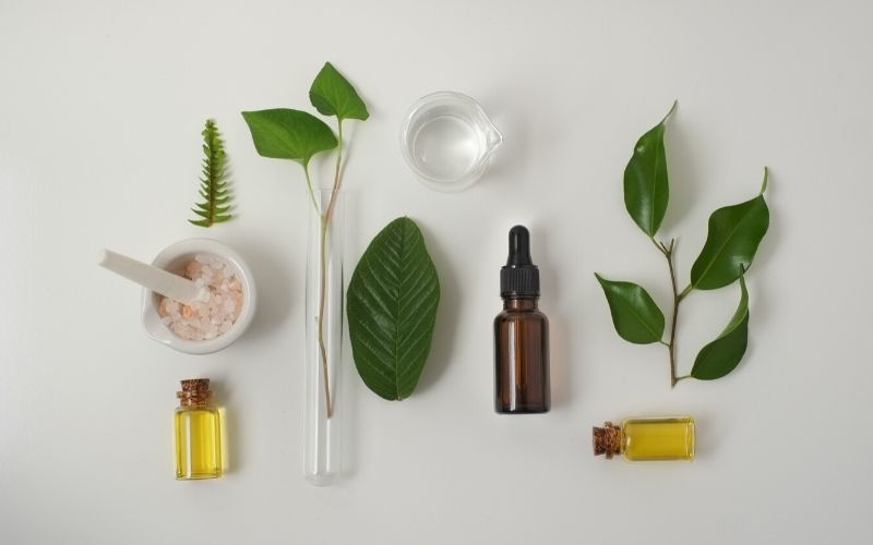 Auf einem weißen Tisch liegen kleine Fläschchen in braun und durchsichtig mit gelber öliger Flüssigkeit im Inneren. Daneben sind ein Schälchen mit groben rosafarbenen Salz und unterschiedliche grüne Blätter und Pflanzen angeordnet.