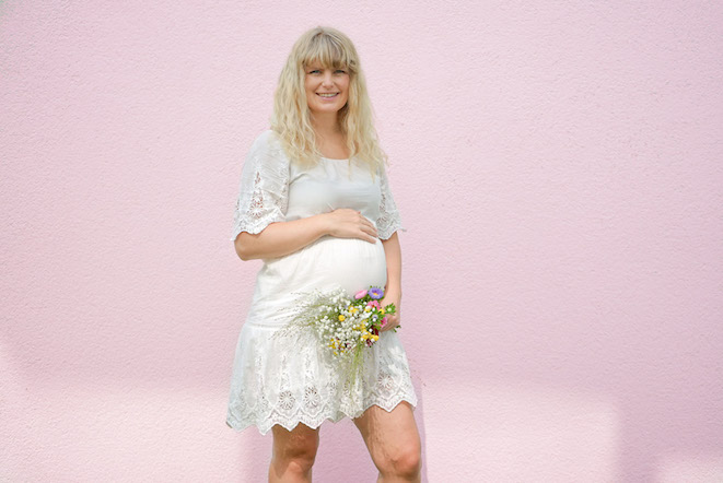 blonde-schwangere-frau-steht-vor-rosa-wand-lacht-haelt-den-bauch