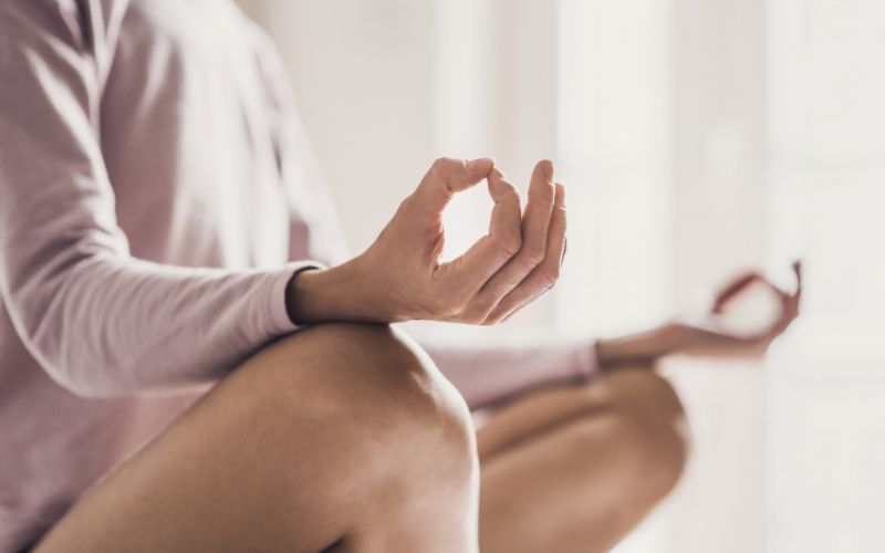 Frau mit rosafarbenem Oberteil sitzt im Schneidersitz, einer Meditationspose und hält ihre Hände zu einem Mudra – Daumen und Zeigefinger berühren sich jeweils – auf ihren Knien.