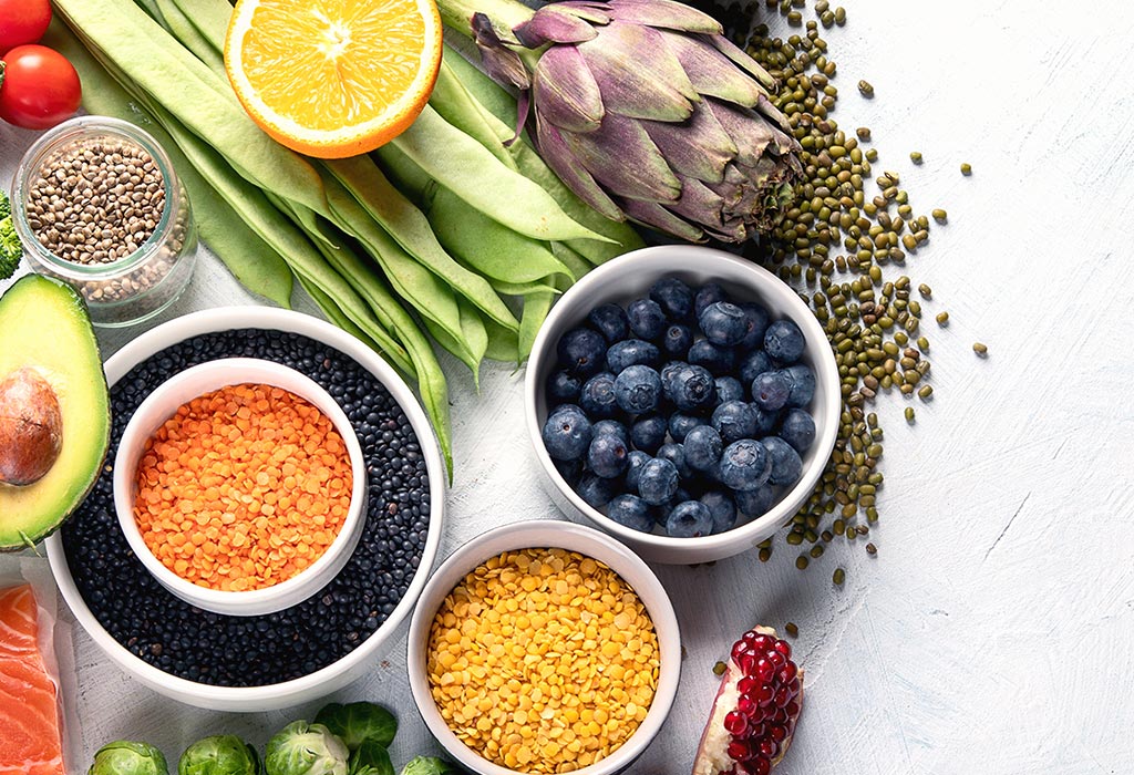 mehrere Schälchen gefüllt mit unterschiedlichen Sachen wie Heidelbeeren, roten und gelben Linsen, Avocado und Mungbohnen stehen nebeneinander auf einem weißen Untergrund.