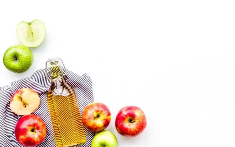 Glasflasche mit gelblicher Flüssigkeit (Apfelessig) liegt auf einem gestreiften Küchenhandtuch. Daneben sind Äpfel in unterschiedlichen Farben zu sehen. Der Hintergrund ist weiß.