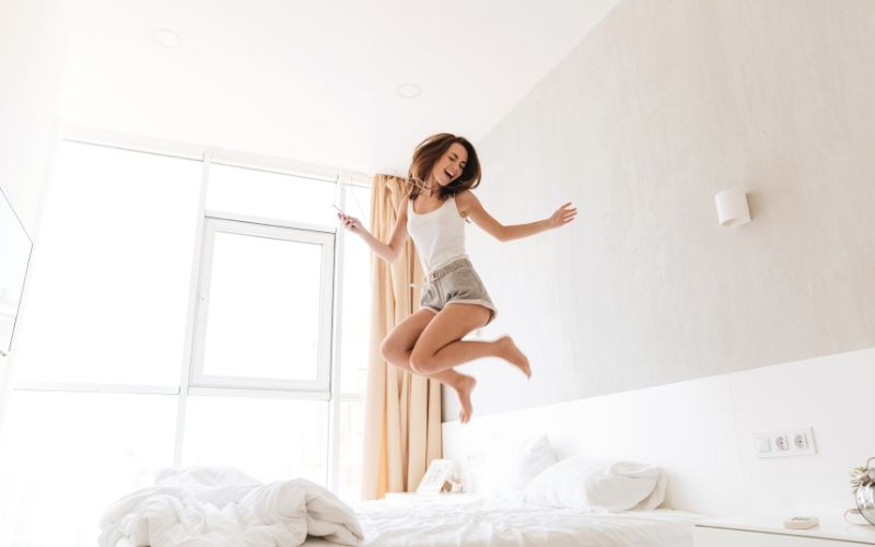 Fröhlich aussehende Frau springt auf dem Bett mit weißen Bettlaken.