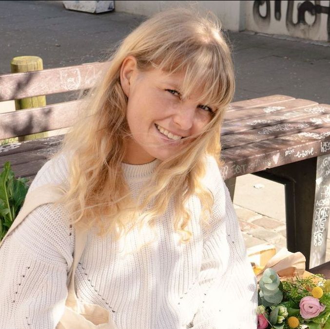 Julia von InnoNature, eine blonde Frau mit hellem Strickpullover, sitzt auf einer Bank im Freien und lächelt in die Kamera.
