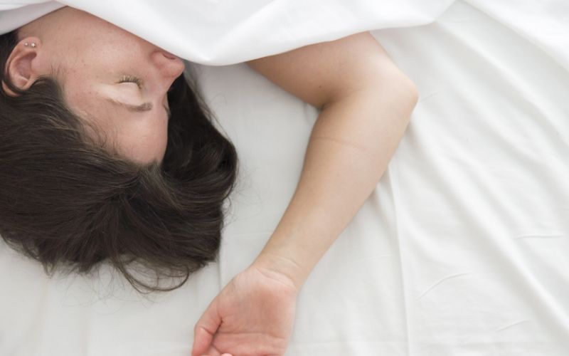 Frau liegt unter einer Decke, zu sehen ist nur ihr Gesicht, die dunklen Haare und ein Arm, den sie halb nach oben hin ausstreckt. Die Augen sind geschlossen.