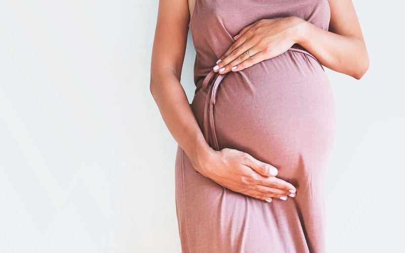 Schwangere Frau mit rosa Kleid hält sich den Bauch. Kopf und Beine der Frau sind nicht zu sehen.