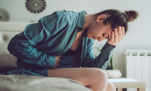junge-frau-sitzt-auf-dem-bett-und-haelt-sich-kopf-und-bauch-als-haette-sie-schmerzen