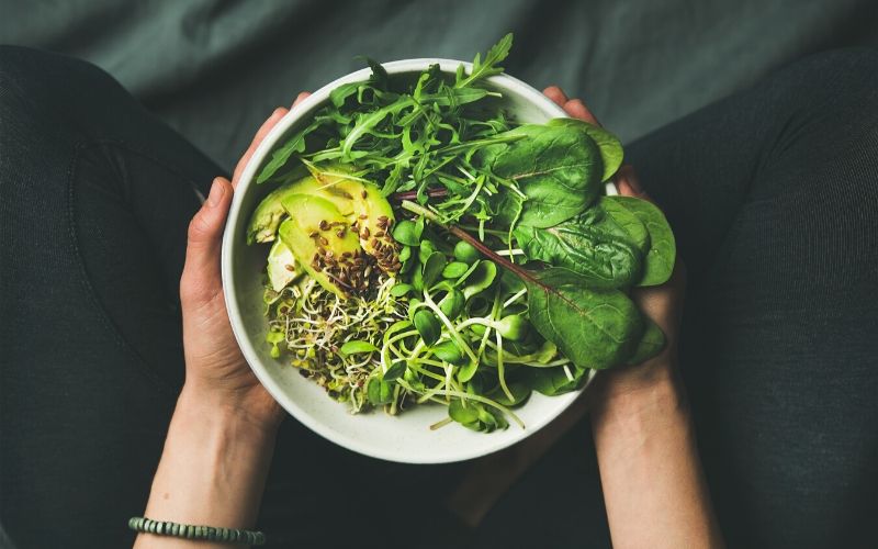 Weibliche Hände halten eine weiße Schale mit unterschiedlichen grünem Blattgemüse darin. Außerdem sind Avocado, Leinsamen und Sprossen zu sehen.