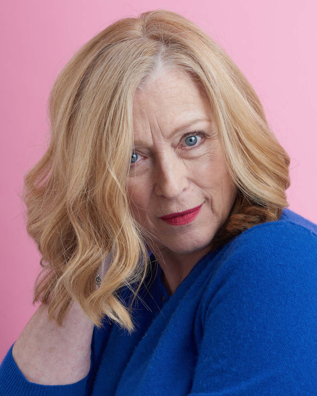 A woman in a blue sweater posing against a pink background - Josh Wood Hair Colour Expert