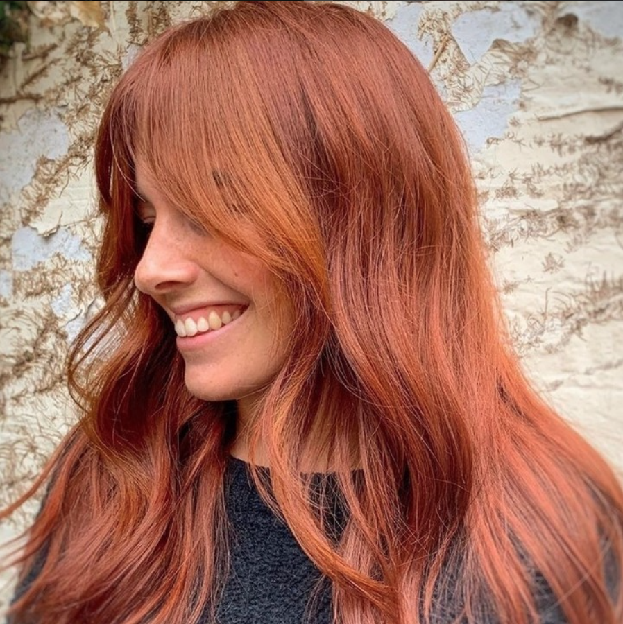 A woman with long red hair smiling in front of a wall - Josh Wood Hair Colour Expert