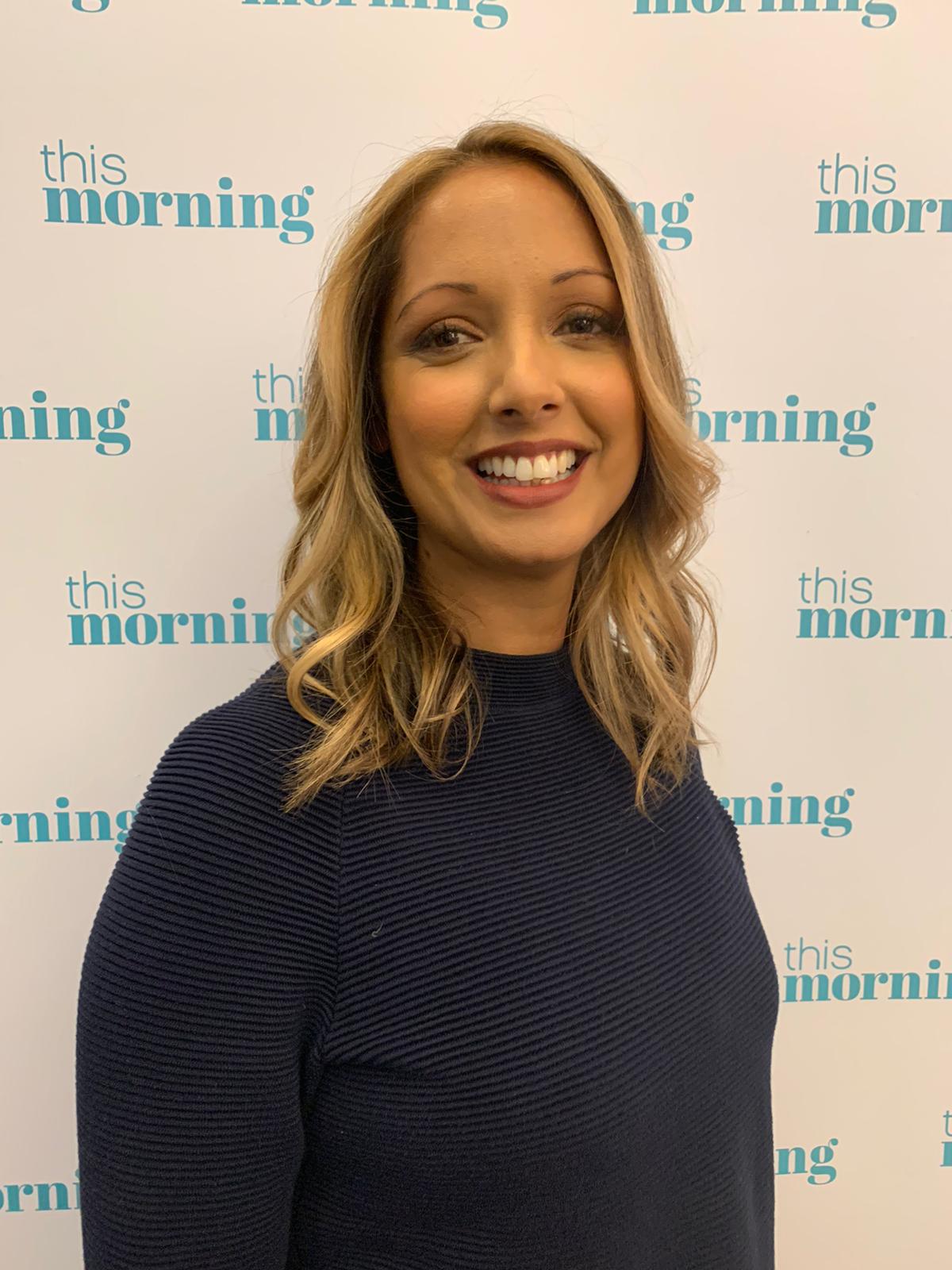 A woman in a blue shirt is smiling in front of a wall - Josh Wood Hair Colour Expert